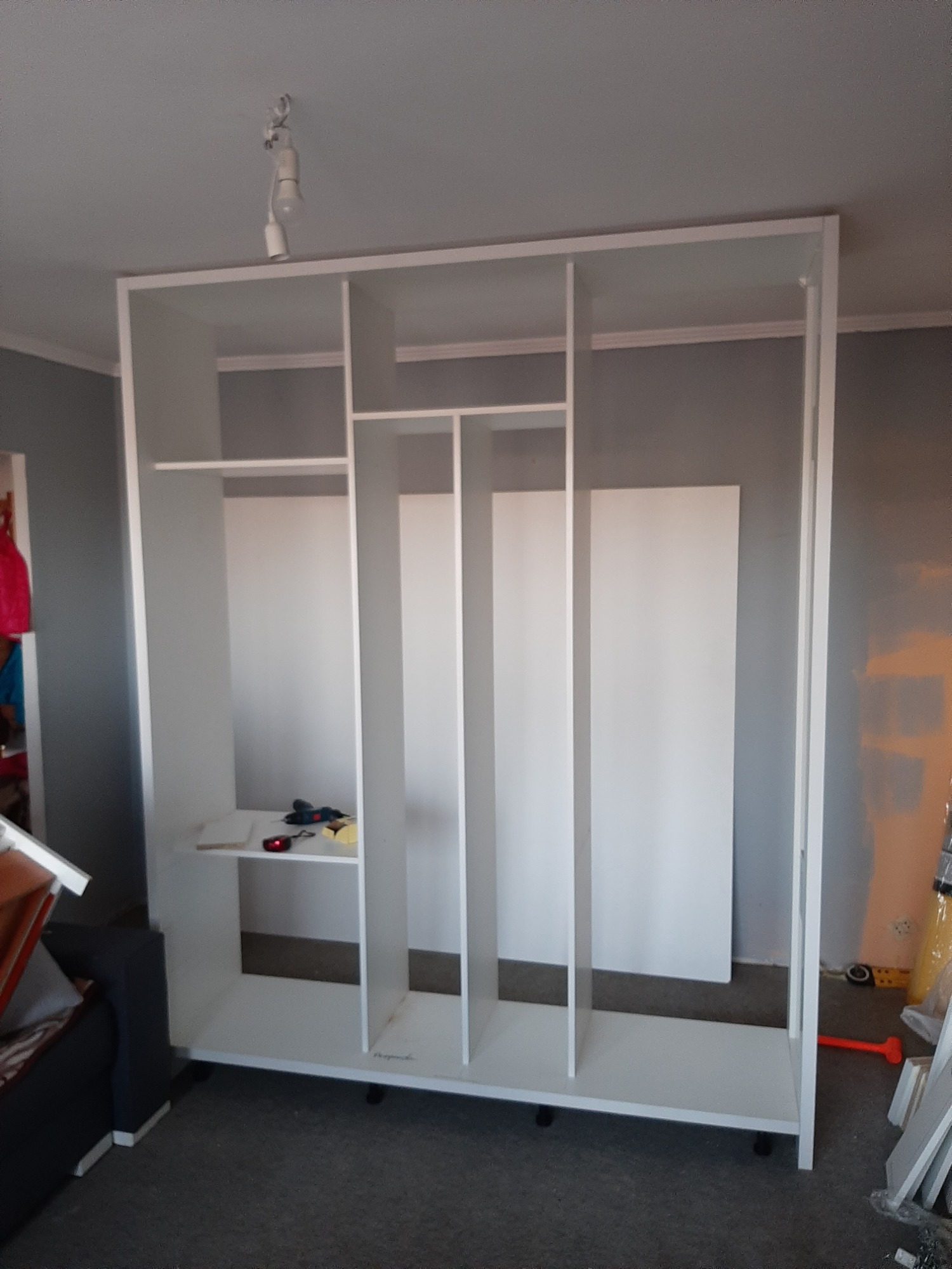 Wnętrze szafy w trakcie montażu z widocznymi narzędziami na półce, surowa konstrukcja z białych płyt MDF, szara ściana w tle.