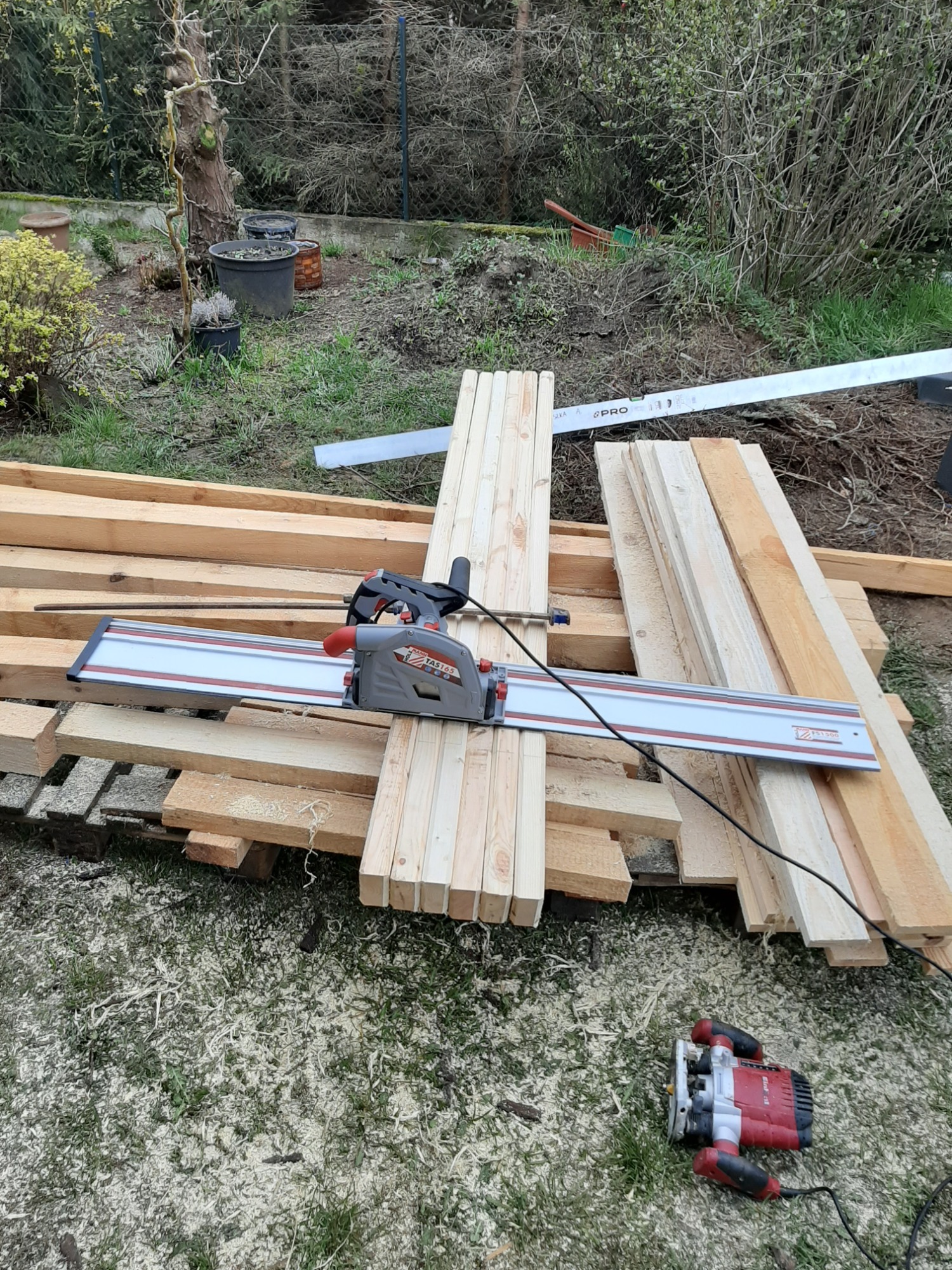 Przygotowane deski ułożone na trawie, na nich pilarka zagłębiarka z szyną prowadzącą i frezarka, w tle ogród.