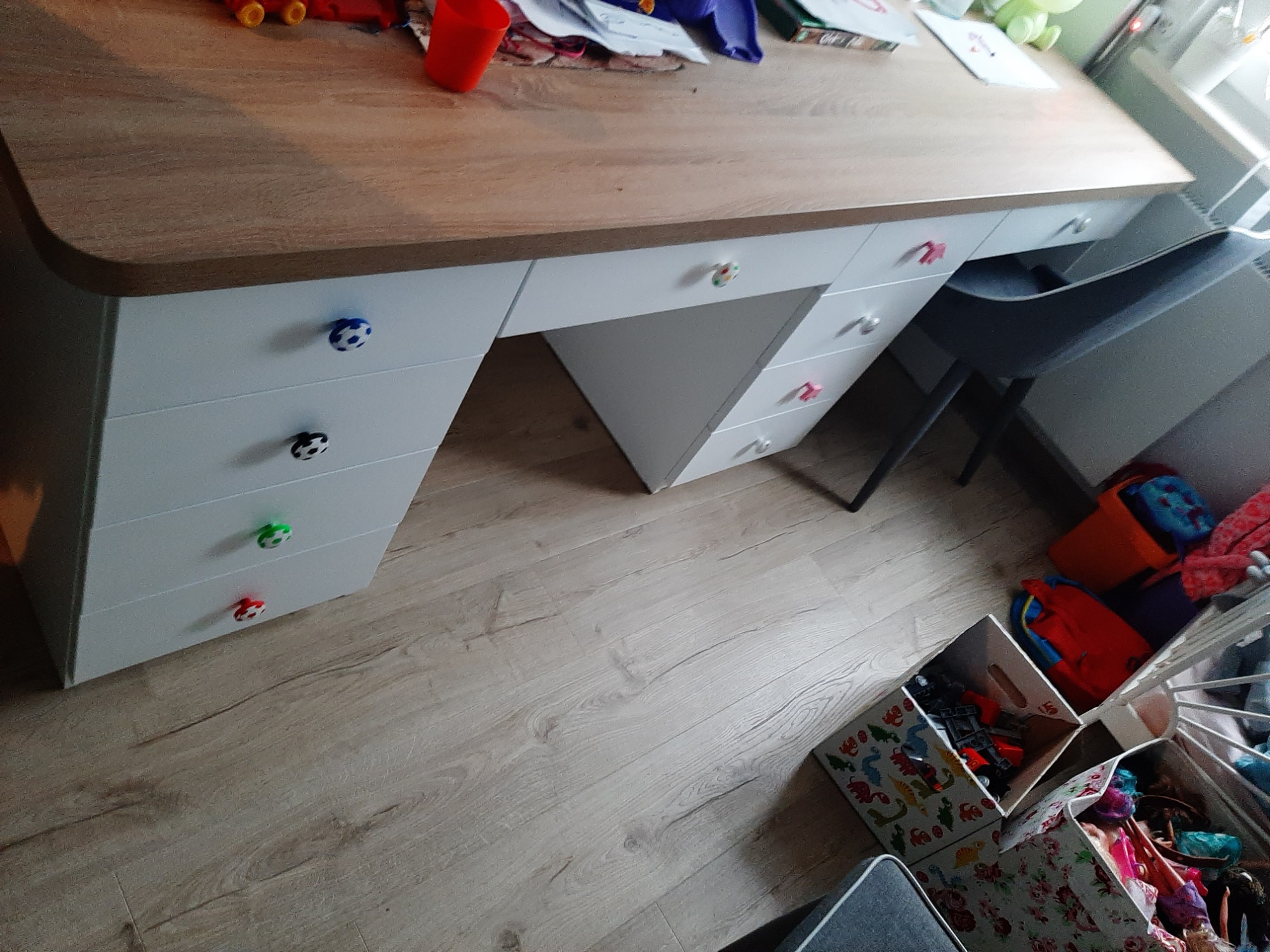 Biurko dziecięce z blatem imitującym drewno i białymi szafkami z szufladami, których uchwyty mają kształt piłek do różnych sportów, na blacie zabawki i przybory szkolne, obok pojemniki z zabawkami.