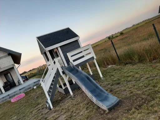 Nowoczesny plac zabaw w formie domku ze zjeżdżalnią, pomalowany na szaro i biało, na tle łąki i ogrodzenia, obok domu jednorodzinnego, widoczne zabawki na trawie.