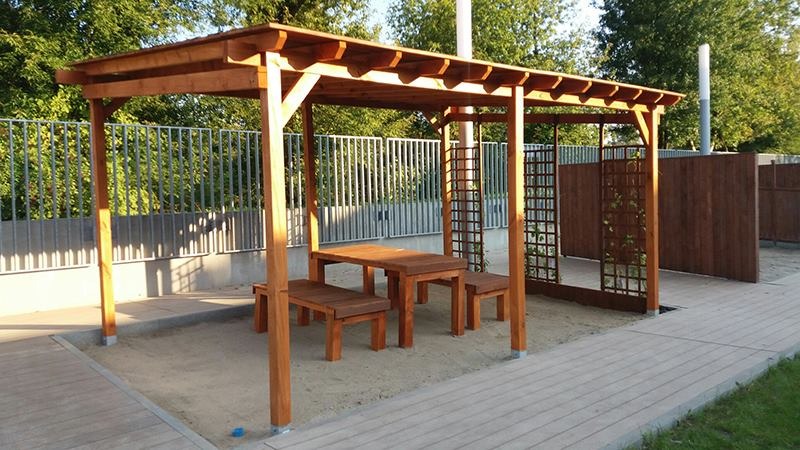 Drewniana altana z prostokątnym stołem i dwoma ławkami, umieszczona na piaskowym podłożu obok chodnika, z metalowym ogrodzeniem w tle.