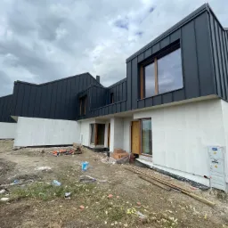 Nowoczesny budynek z grafitową, pionowo układaną blachą na dachu i elewacją w trakcie wykańczania, widoczny bałagan na budowie i pochmurne niebo.