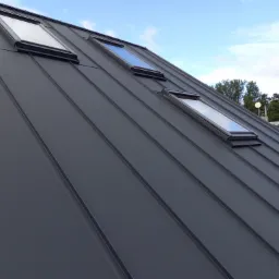 Nowoczesny, grafitowy dach z blachy na rąbek stojący z trzema oknami dachowymi, widok z dołu na tle błękitnego nieba.