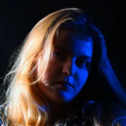 Portret młodej kobiety z blond włosami oświetlonej niebieskim światłem na ciemnym tle.