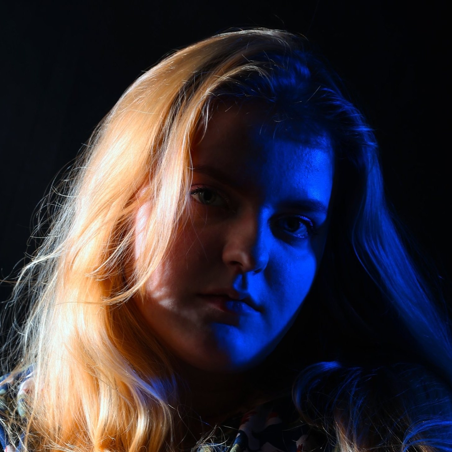 Portret młodej kobiety z blond włosami oświetlonej niebieskim światłem na ciemnym tle.