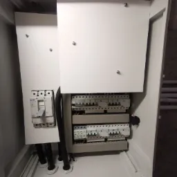 Otwarta skrzynka rozdzielcza z zamontowanymi bezpiecznikami i wyłącznikiem głównym Eaton, widoczne kable wchodzące do skrzynki przez dławiki.