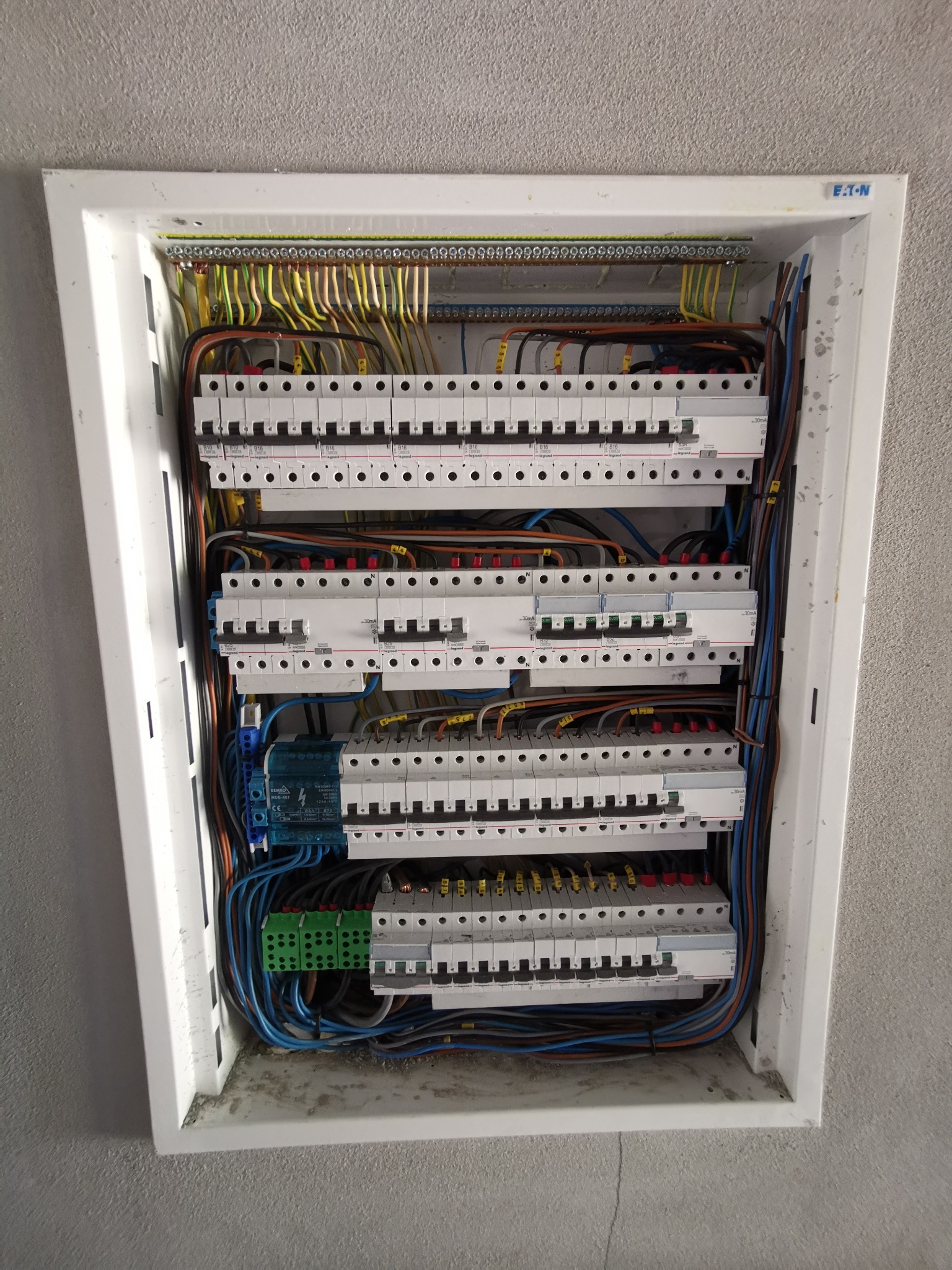 Rozdzielnica elektryczna z widocznymi bezpiecznikami, przewodami i listwami zaciskowymi, zamontowana w białej obudowie w szarej ścianie. Widoczne oznaczenia producentów bezpieczników: Legrand...