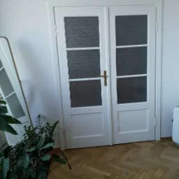 Białe, dwuskrzydłowe drzwi z czterema szybkami o fakturze piaskowanego szkła, z mosiężną klamką, na tle parkietu ułożonego w jodełkę, z rośliną doniczkową w lewym dolnym rogu i lustrem opartym...