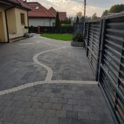 Nowoczesny podjazd z ciemnoszarej kostki brukowej z dekoracyjnymi wstawkami z jasnej kostki, prowadzący do domu z czerwonym dachem i metalowej bramy.