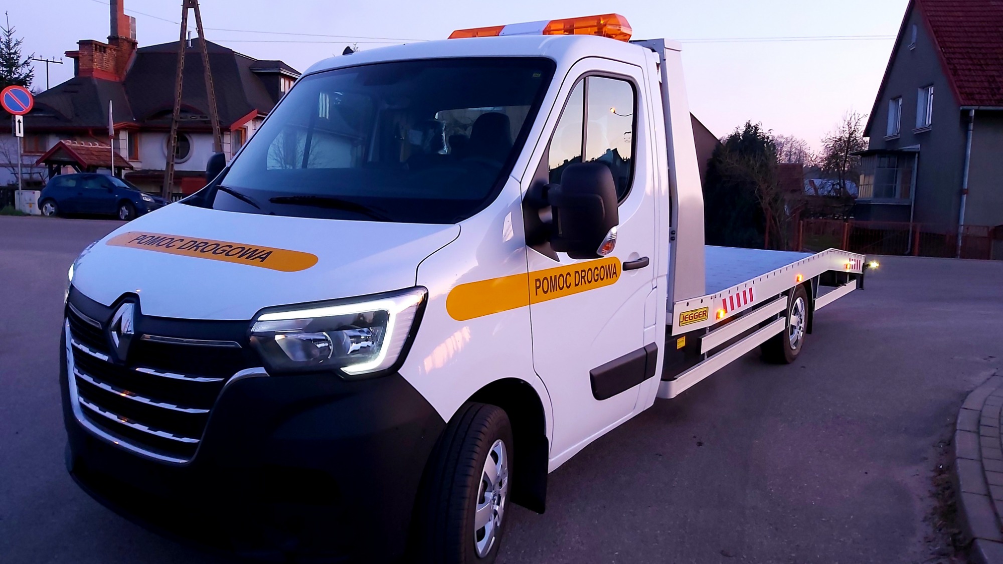 Biała laweta Renault Master z napisem 'Pomoc Drogowa' na żółtym tle, zaparkowana przy drodze w scenerii zmierzchu. Widoczne włączone światła ostrzegawcze na dachu i przednie reflektory.