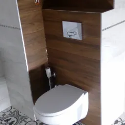 Wisząca toaleta z chromowanym przyciskiem spłukującym, drewnopodobne panele ścienne i wzorzysta podłoga w odcieniach szarości i bieli.