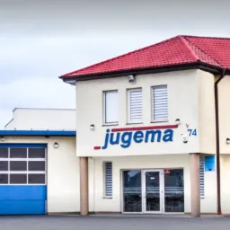 Budynek firmy Jugema 74 z czerwonym dachem, niebieską bramą garażową i niebieskim logo na fasadzie, otoczony drzewami.
