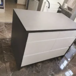 Szara komoda z trzema białymi, lakierowanymi szufladami, stojąca na ciemnej marmurowej podłodze, widoczna z boku pod kątem.