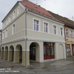 Odnowiona kamienica z podcieniami i nową elewacją w odcieniach beżu, z widoczną częścią dachu z czerwonej dachówki i białymi oknami. Na pierwszym planie brukowana ulica i rzeźba. Widoczne...