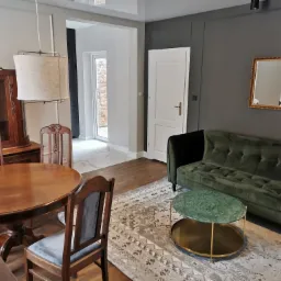 Elegancki salon z drewnianym stołem, krzesłami, witryną, zieloną sofą i okrągłym stolikiem kawowym na wzorzystym dywanie. Lustro w złotej ramie na szarej ścianie.