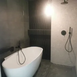 Nowoczesna łazienka z wolnostojącą białą wanną, czarną armaturą i grafitową ścianą wyłożoną płytkami w kształcie rombów, oświetlona górnym światłem.