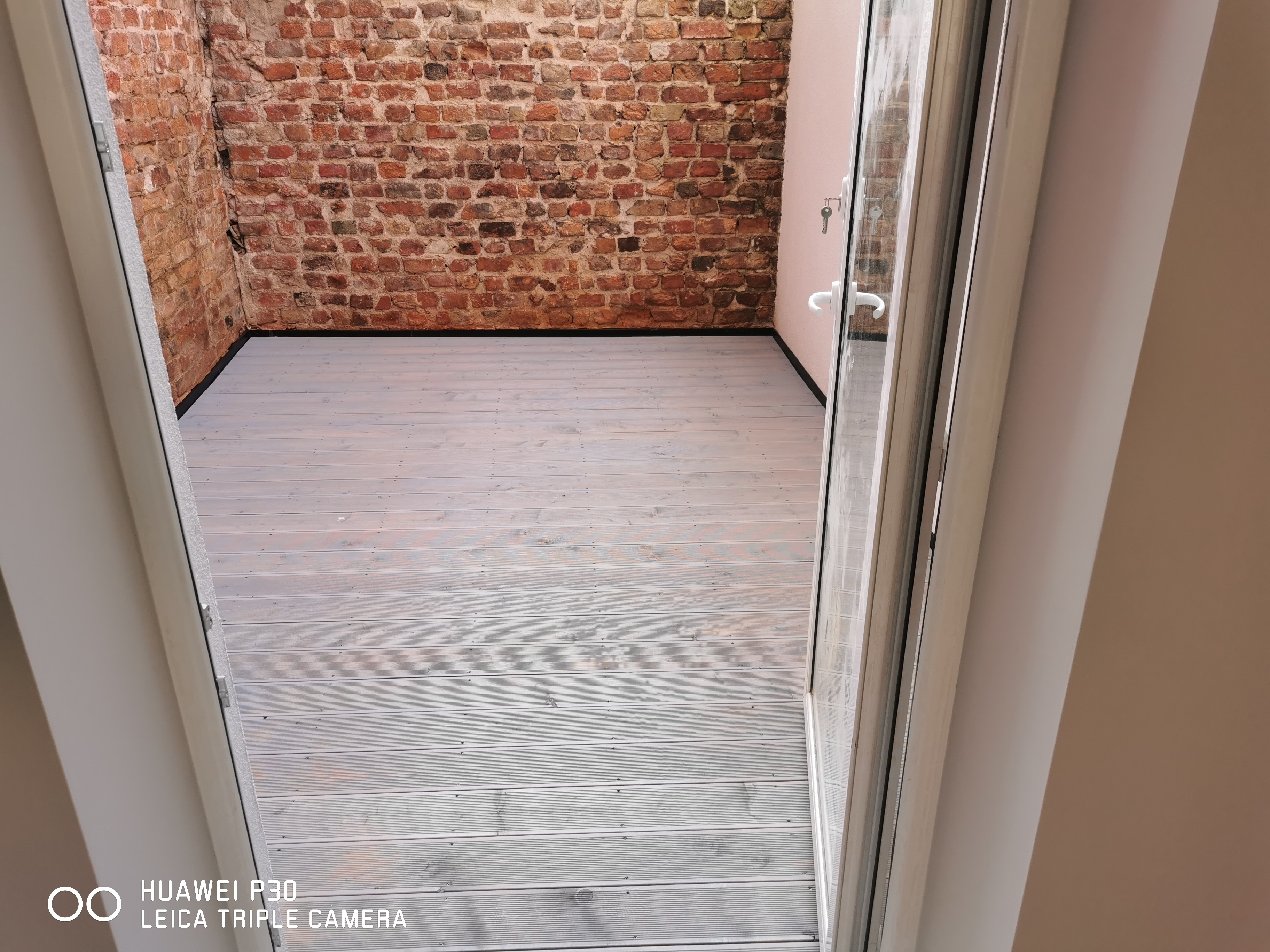 Szare deski tarasowe ułożone na zewnątrz, widoczne zza otwartych drzwi balkonowych, z ceglaną ścianą w tle.