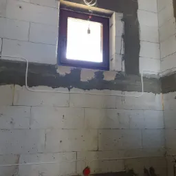 Wewnętrzna ściana z betonu komórkowego w trakcie prac wykończeniowych, z widocznym oknem, przewodami elektrycznymi ułożonymi na ścianie i puszką instalacyjną w kolorze czerwonym przy podłodze.
