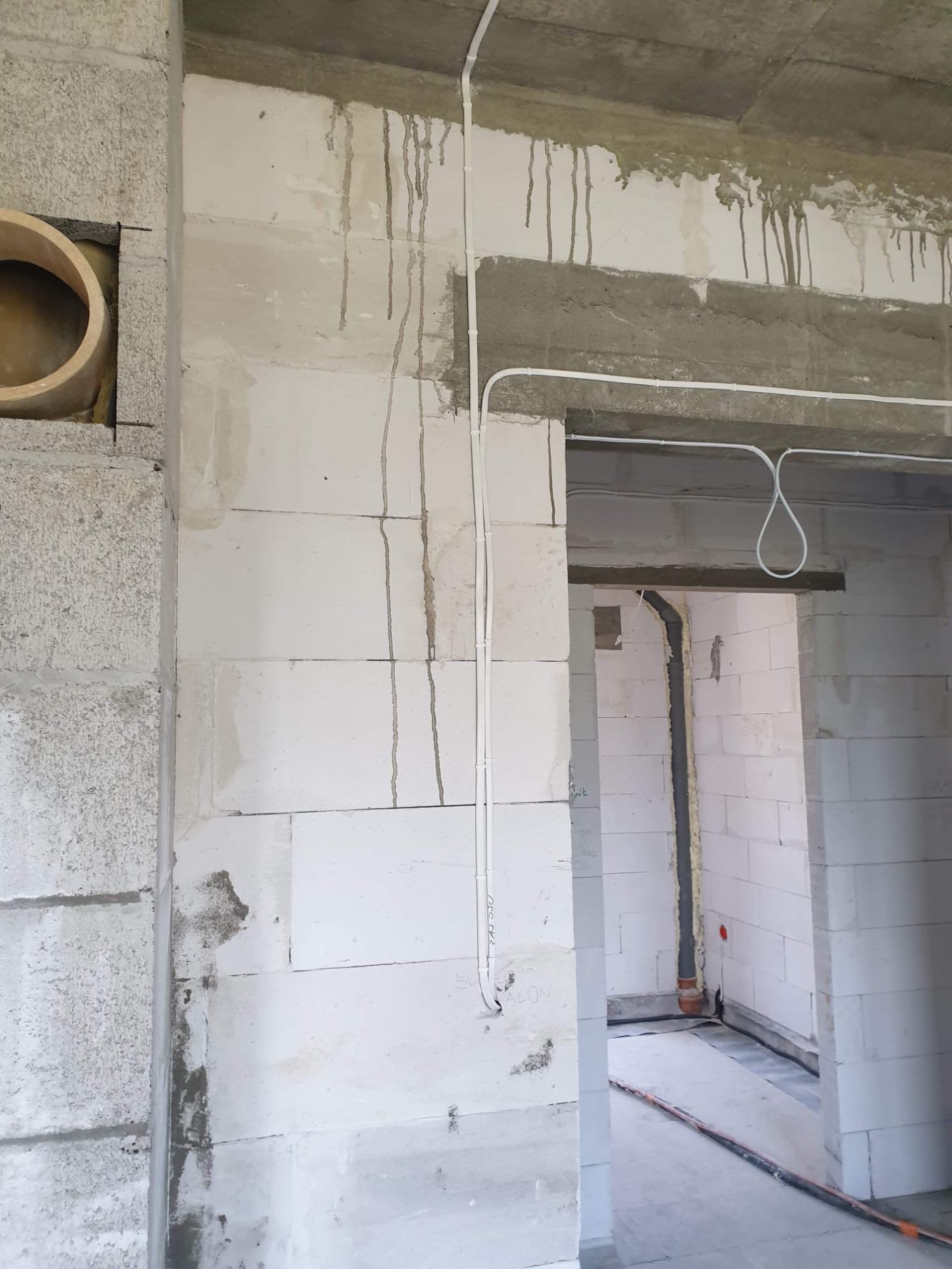 Surowy stan wnętrza w trakcie prac wykończeniowych: widoczne białe ściany z betonu komórkowego, wystające przewody elektryczne poprowadzone po ścianie oraz otwór wentylacyjny z drewnianym...