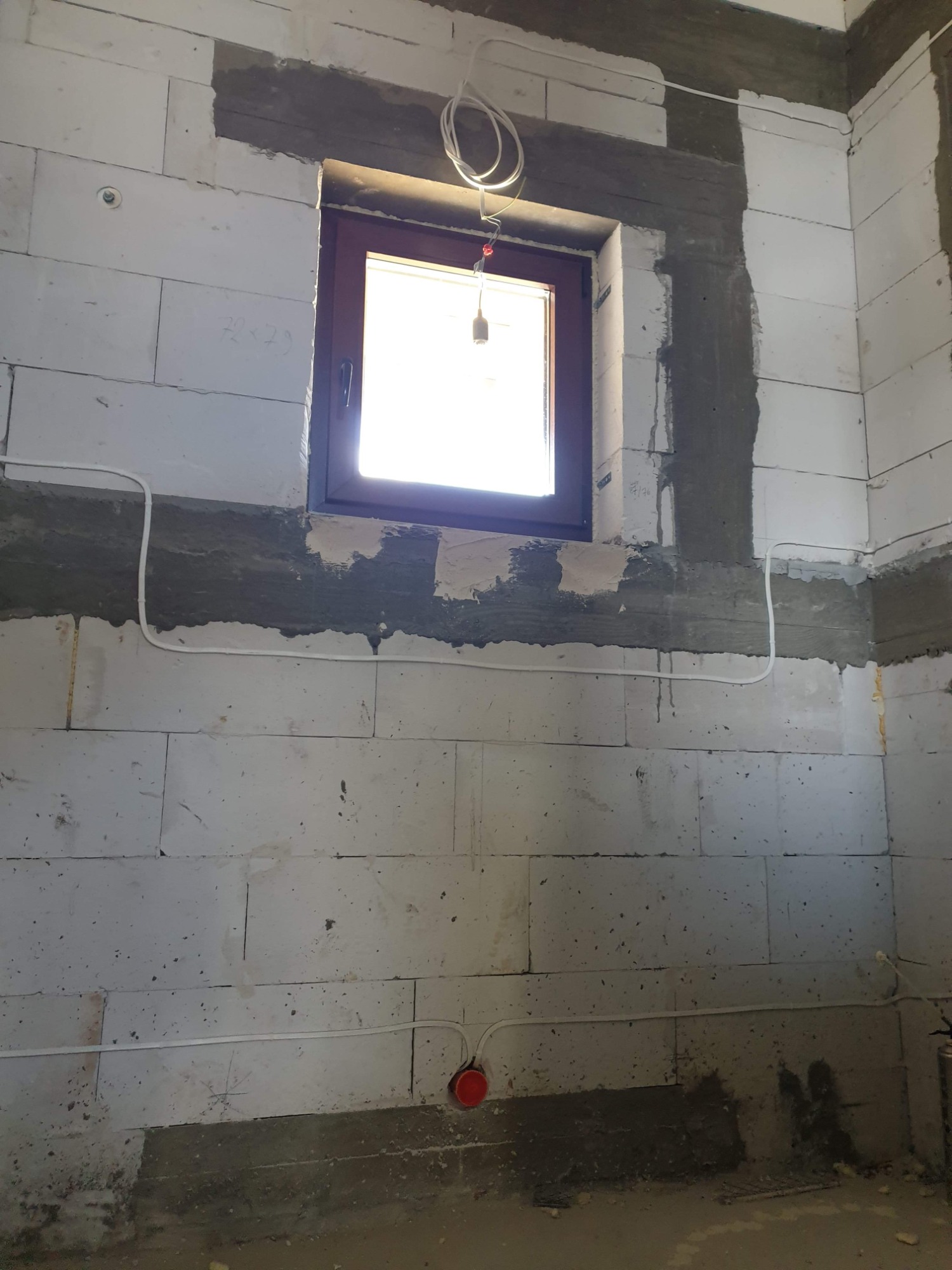 Wewnętrzna ściana z betonu komórkowego w trakcie prac wykończeniowych, z widocznym oknem, przewodami elektrycznymi ułożonymi na ścianie i puszką instalacyjną w kolorze czerwonym przy podłodze.