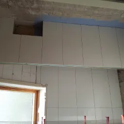 Układanie białych płytek ściennych w łazience, widoczne niedokończone ściany i sufit, okno z drewnianą ramą i plastikowe kliny poziomujące płytki.