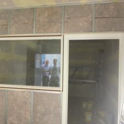 A2 Przedsiebiorstwo Ogólnobudowlane (AIDA-GP) - Montaż okna i drzwi w pomieszczeniu z izolacją ścian, widoczne profile metalowe i wełna mineralna, w tle odbicie dwóch osób i drabina.