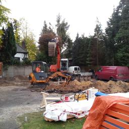 A2 Przedsiebiorstwo Ogólnobudowlane (AIDA-GP) - Dwie koparki przygotowują teren pod budowę zadaszenia dla samochodów, widoczne materiały budowlane i narzędzia DeWalt, w tle zaparkowane busy i przyczepa.