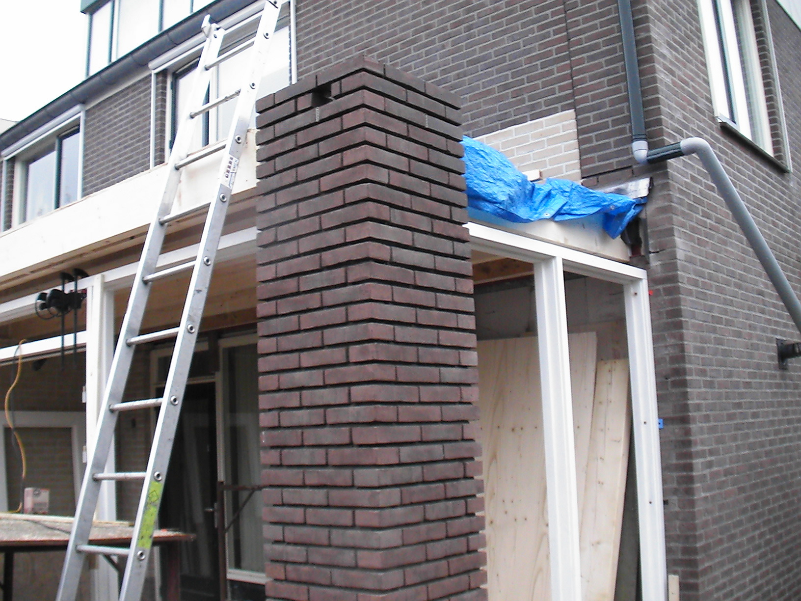 Przybudowa domu w trakcie prac: widoczna murowana kolumna z cegły, drabina oparta o ścianę, zabezpieczenie folią budowlaną, nowa rama okienna w surowym stanie.