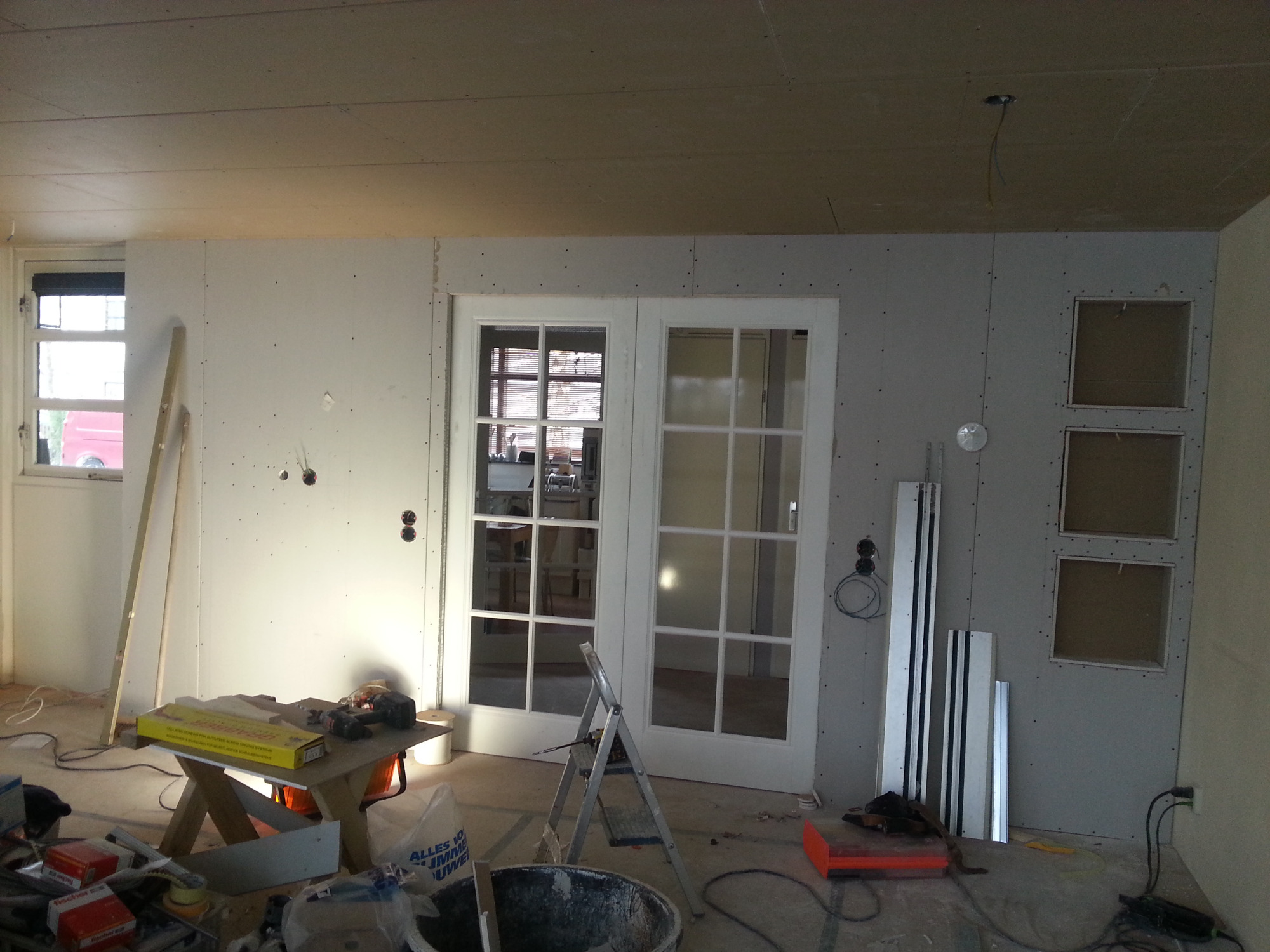 Wnętrze w trakcie remontu: zamontowana ścianka działowa z podwójnymi, przeszklonymi drzwiami przesuwnymi, widoczne narzędzia, drabina i materiały budowlane, ściany pokryte płytami...