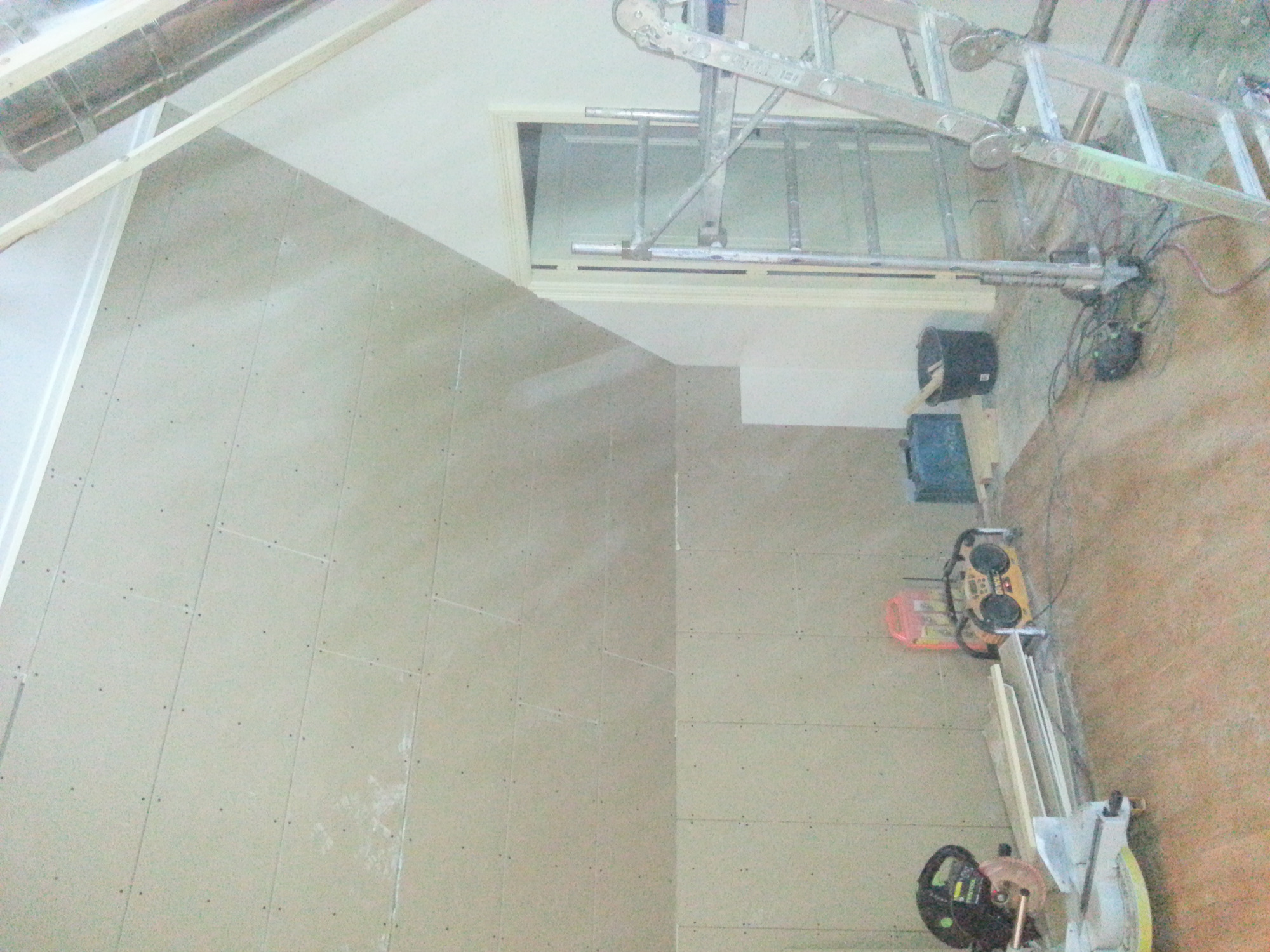 Płyty gipsowo-kartonowe zamontowane na ścianie pod skosem, drabina oparta o ramę okienną, widoczne narzędzia i materiały budowlane.