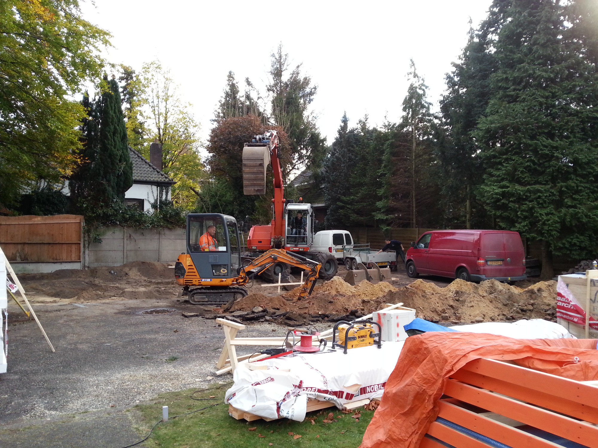 Dwie koparki przygotowują teren pod budowę zadaszenia dla samochodów, widoczne materiały budowlane i narzędzia DeWalt, w tle zaparkowane busy i przyczepa.