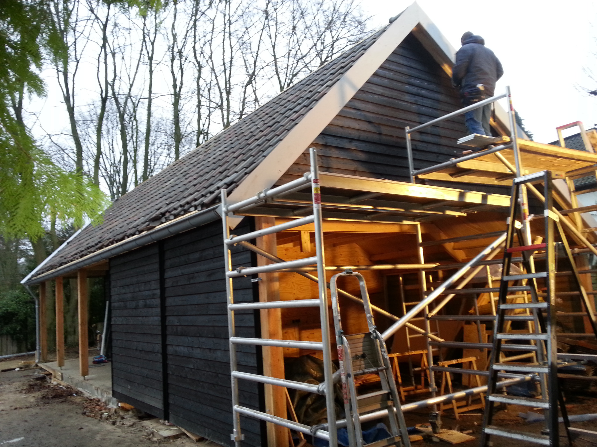 Budowa zadaszenia pod auta w stylu atelier: czarne drewniane ściany, dach z dachówki, rusztowanie, pracownik na wysokości.