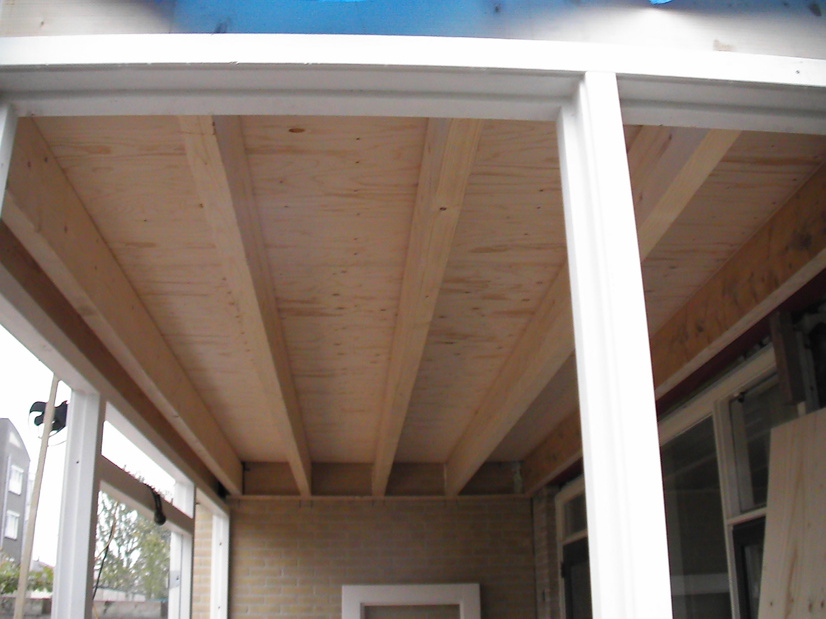Konstrukcja drewnianego stropu przybudówki z widocznymi belkami i deskowaniem, białe ramy okienne w tle, ceglana ściana.