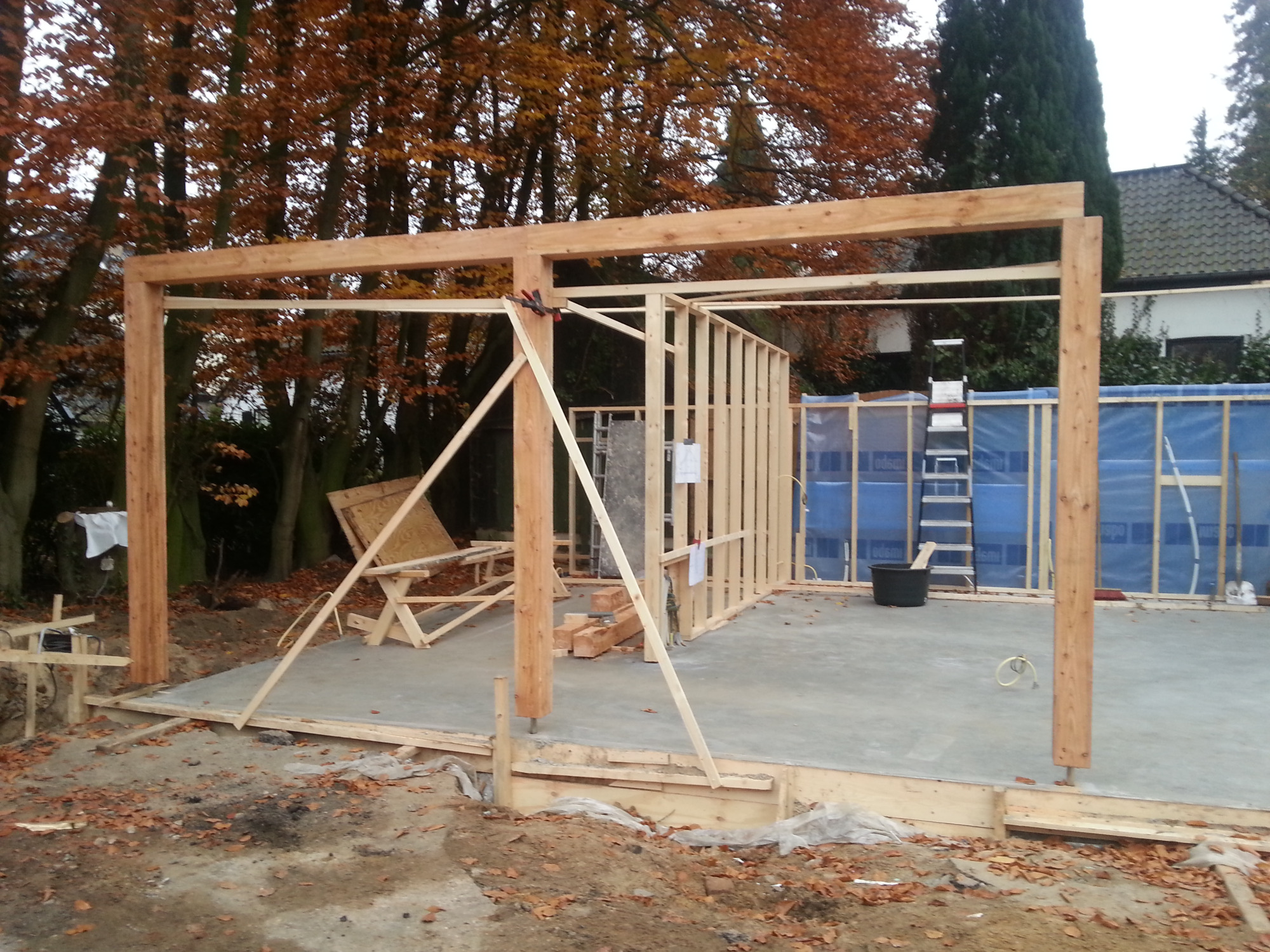 Konstrukcja szkieletowa z drewna, prawdopodobnie zadaszenia, w trakcie budowy na betonowej płycie, jesienne liście w tle.