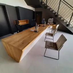 Długi, jasny blat drewniany z białym wykończeniem w nowoczesnym biurze z czarnymi ścianami i schodami. Krzesła tapicerowane w odcieniach szarości.