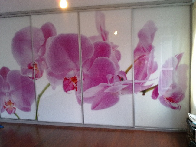 Szafa wnękowa z przesuwnymi drzwiami, na których widnieje duży motyw kwiatowy z różowymi orchideami na białym tle, widoczny fragment pokoju z drewnianą podłogą.