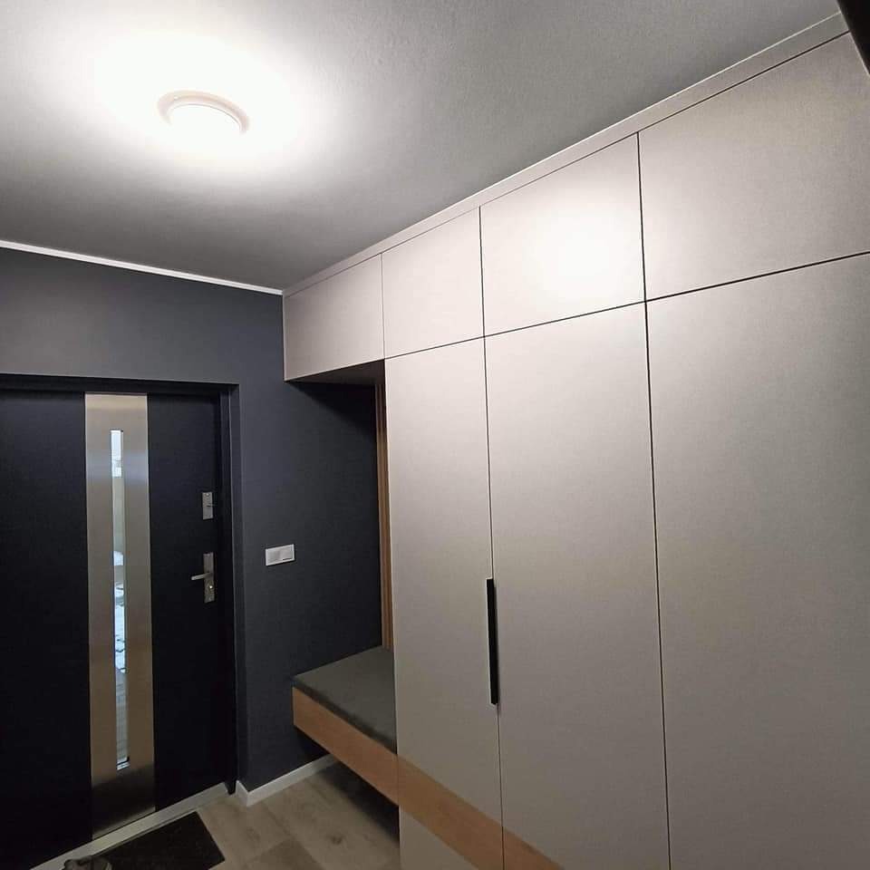 Przestronny przedpokój z zabudowaną szafą z siedziskiem, fronty w kolorze jasnego beżu, czarne, długie uchwyty, ciemnoszara ściana z drzwiami wejściowymi.