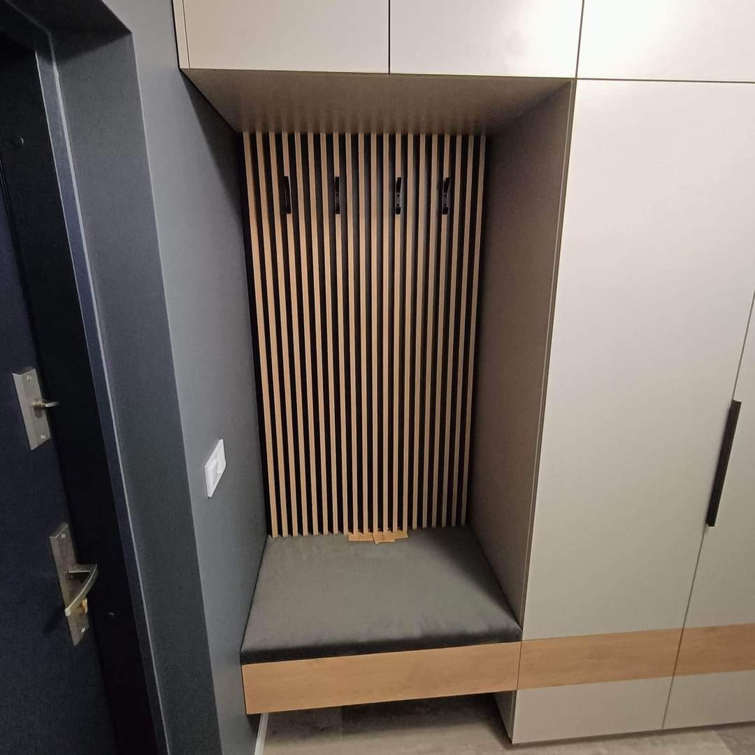 Wnęka z siedziskiem tapicerowanym szarą tkaniną i drewnianymi lamelami na ścianie, wkomponowana w zabudowę szafy w przedpokoju, widoczny fragment drzwi wejściowych.