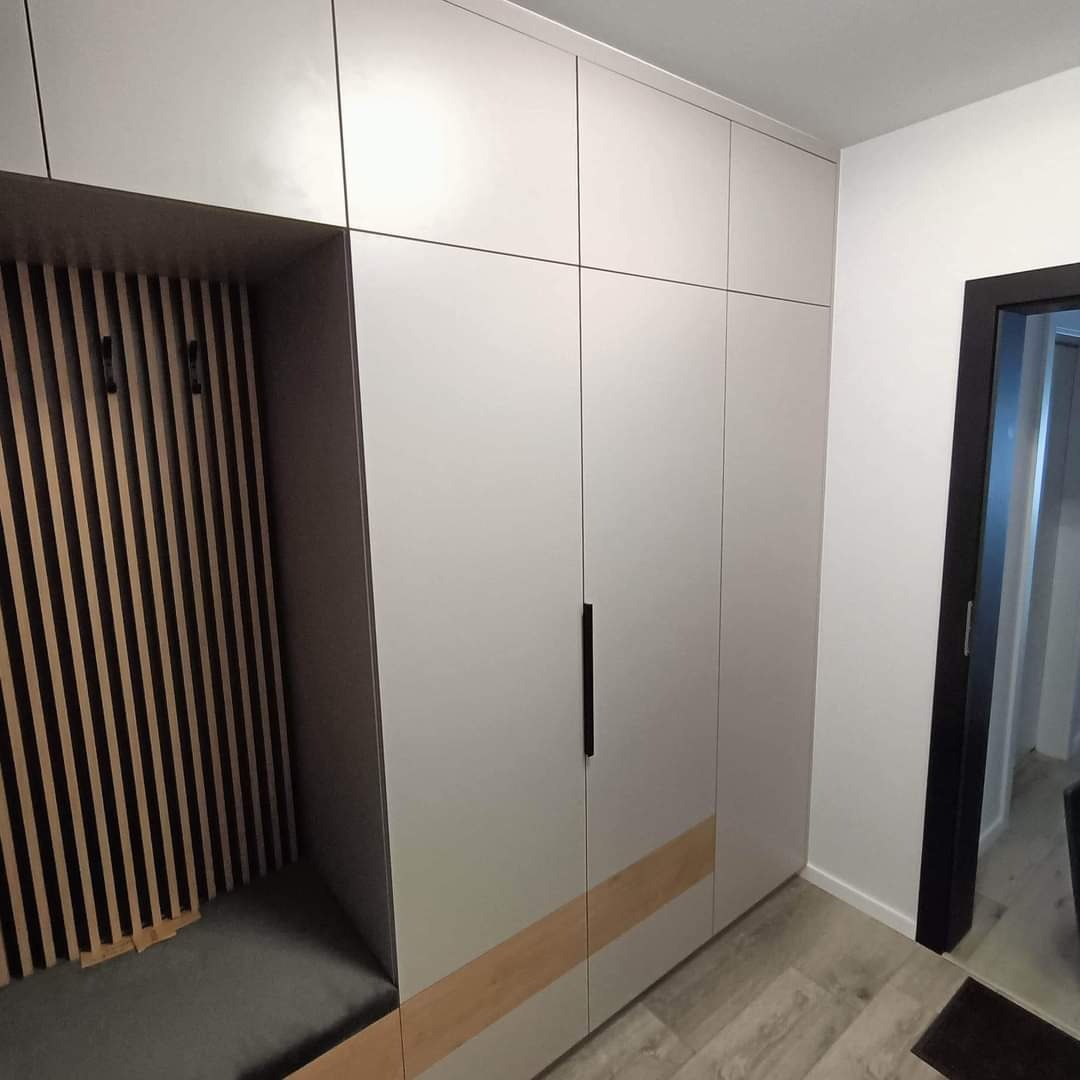 Biała szafa wnękowa z siedziskiem i ażurowym panelem na ścianie w przedpokoju, z czarnymi uchwytami i drewnianym akcentem u dołu, widok na otwarte drzwi.