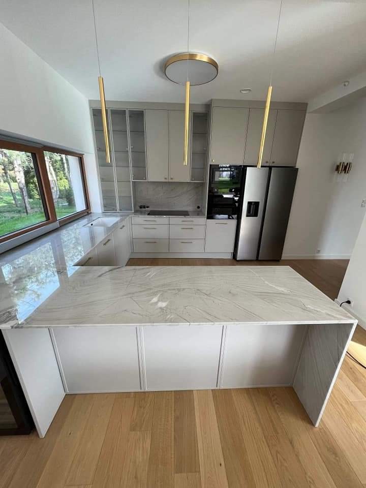 Nowoczesna kuchnia z wyspą, blatem z białego marmuru i złotymi lampami wiszącymi, widok na ogród przez duże okno.