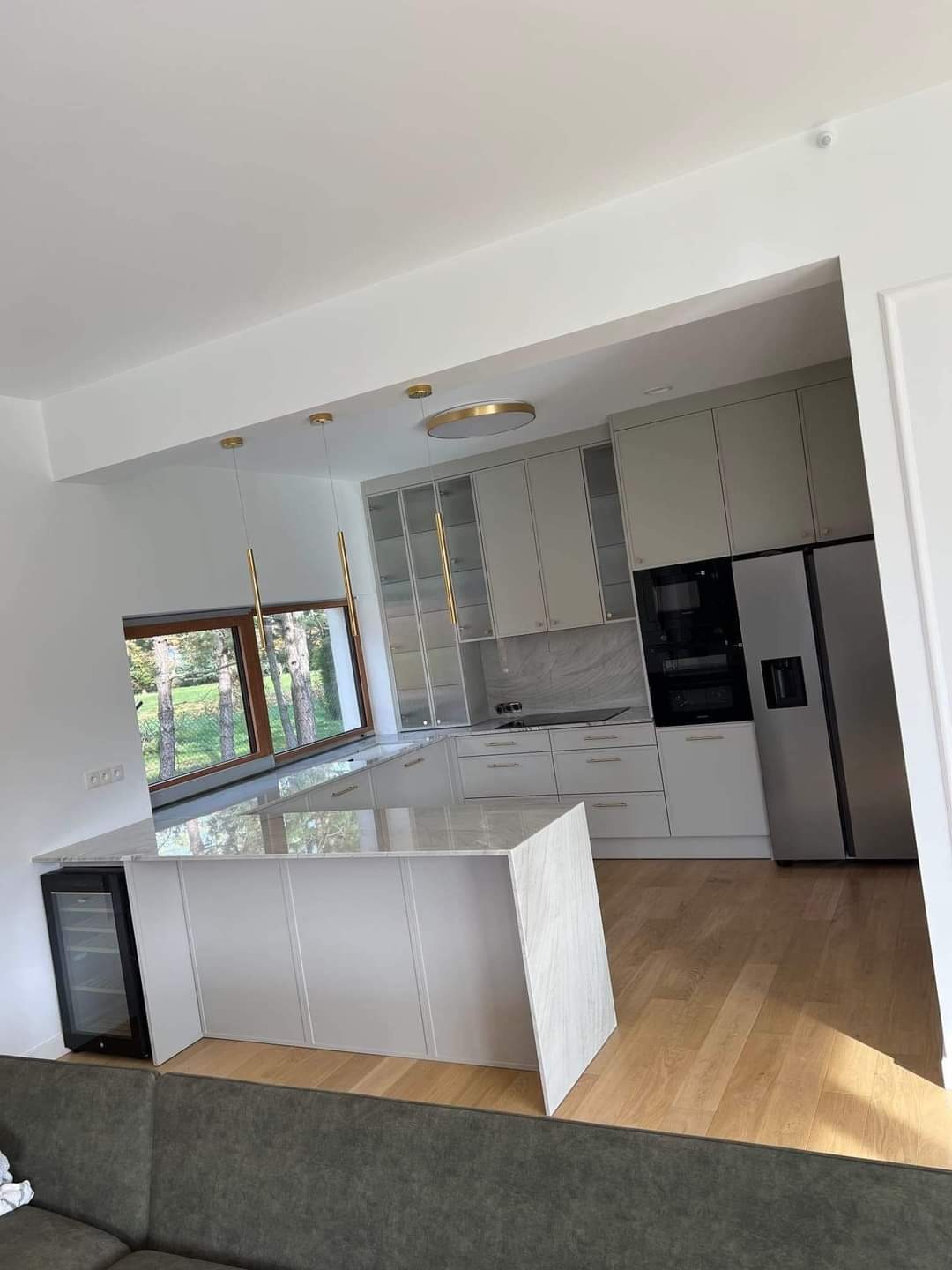 Nowoczesna kuchnia z wyspą i blatem z marmuru, oświetlona złotymi lampami wiszącymi, z widokiem na zieleń za oknem.