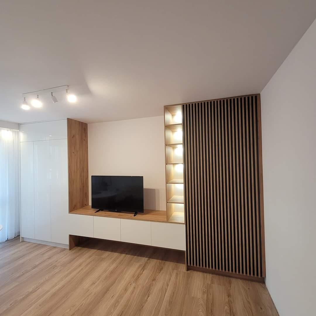 Nowoczesny salon z zabudową meblową: białe szafy, szafka RTV z drewnianym blatem, ażurowa ścianka działowa z podświetlanymi półkami, drewniana podłoga.