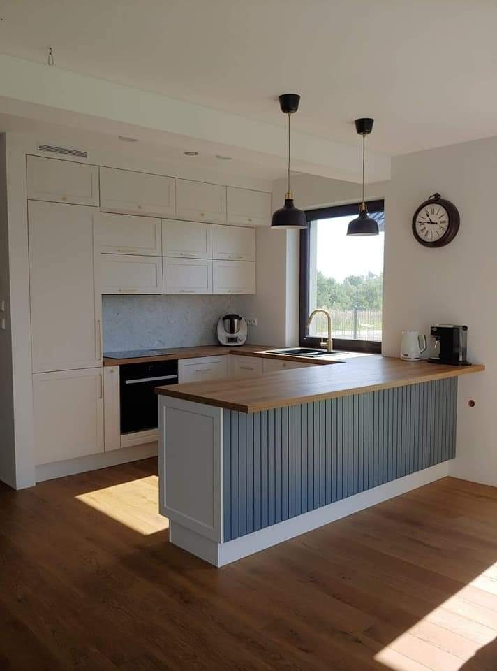 Jasna kuchnia z wyspą, drewnianym blatem i frontami w kolorze szaro-niebieskim. Nad wyspą wiszą dwie czarne lampy. Na ścianie widoczny zegar.