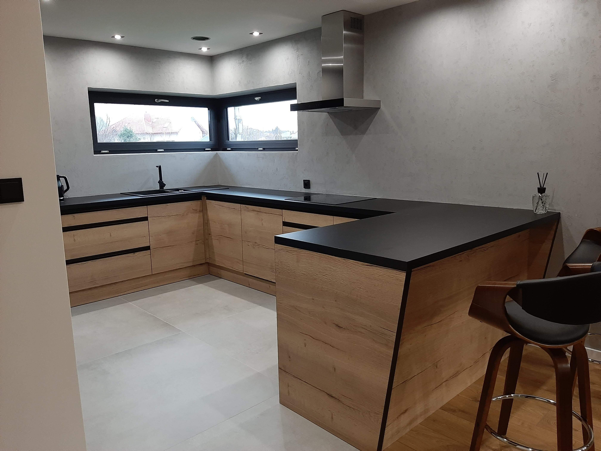 Nowoczesna kuchnia z drewnianymi szafkami i czarnym blatem, widok na okno i minimalistyczny okap.