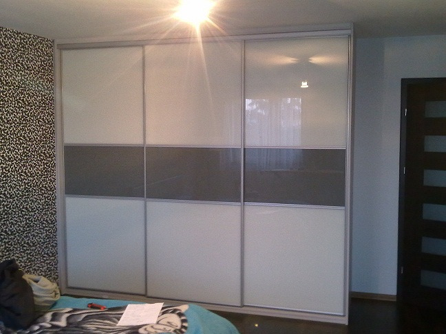 Trzydrzwiowa szafa przesuwna z białymi i szarymi panelami, widoczna w sypialni z wzorzystą tapetą i fragmentem łóżka.