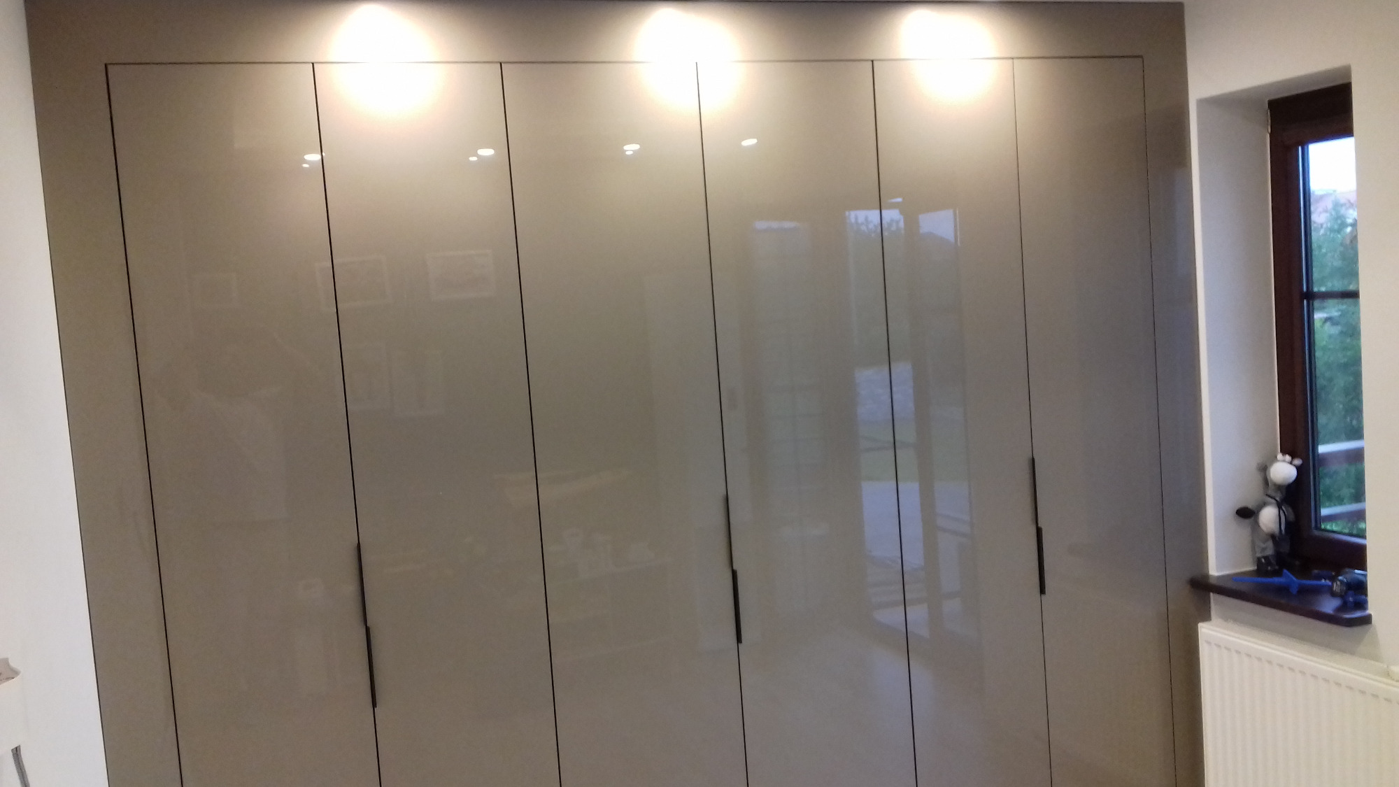 Gładka, beżowa szafa wnękowa z sześcioma drzwiami na wysoki połysk, z minimalistycznymi, czarnymi uchwytami, oświetlona od góry, z odbiciami w drzwiach, stojąca obok okna z dekoracjami na parapecie.