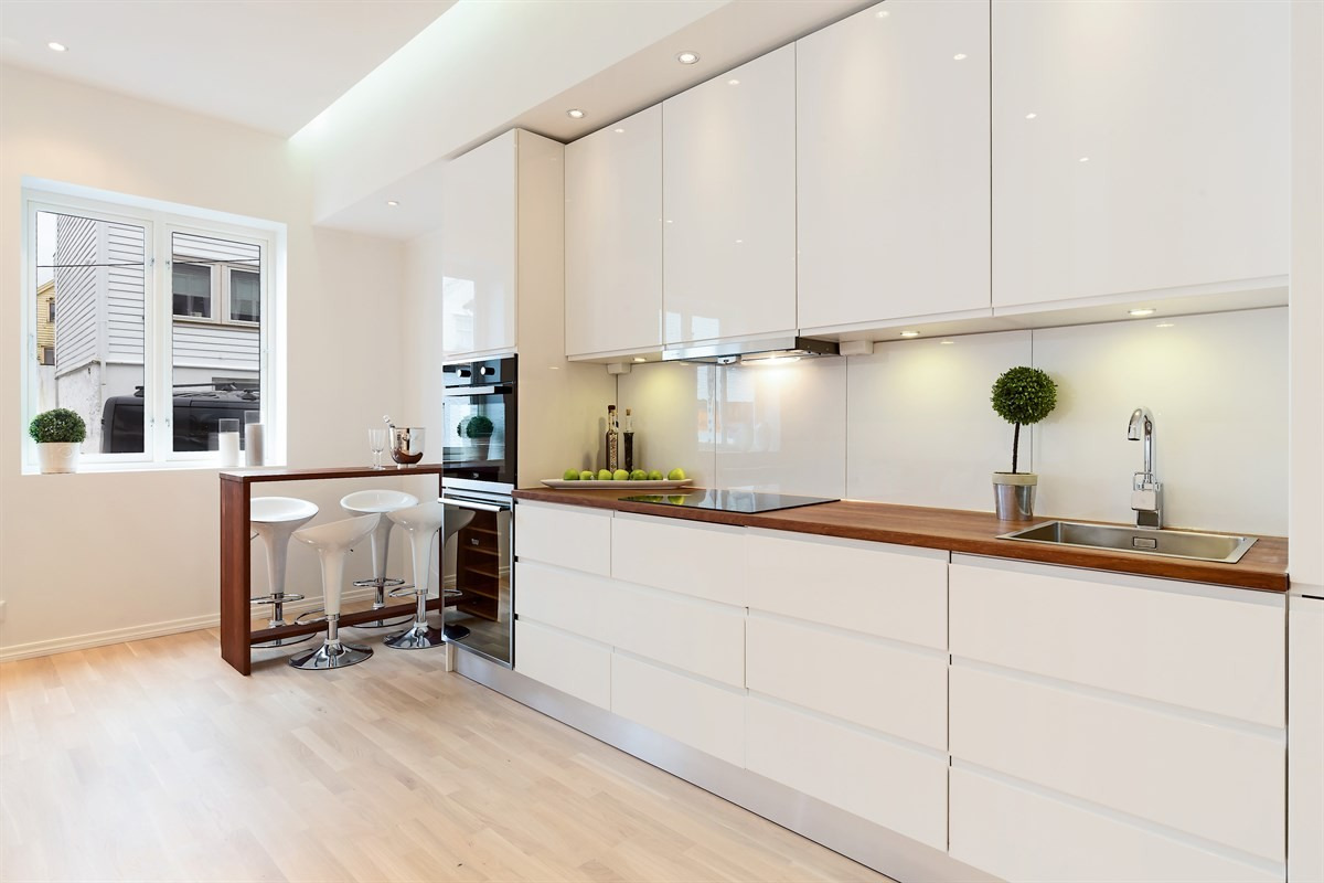 Jasna kuchnia z białymi, lakierowanymi szafkami, drewnianym blatem, barkiem z hokerami i oknem z widokiem na zabudowę jednorodzinną.