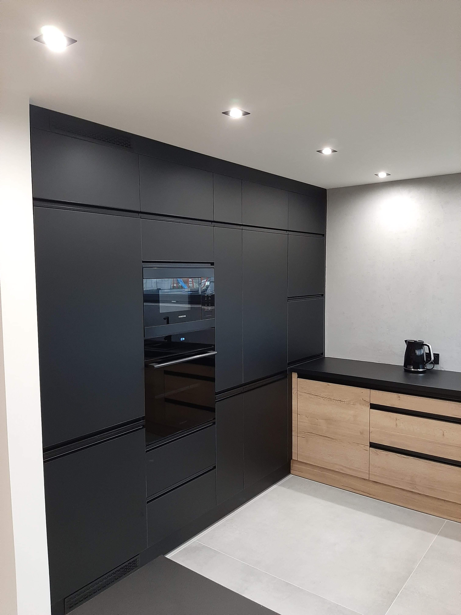 Nowoczesna kuchnia z czarnymi frontami i drewnianymi akcentami, oświetlenie punktowe, czajnik na blacie.