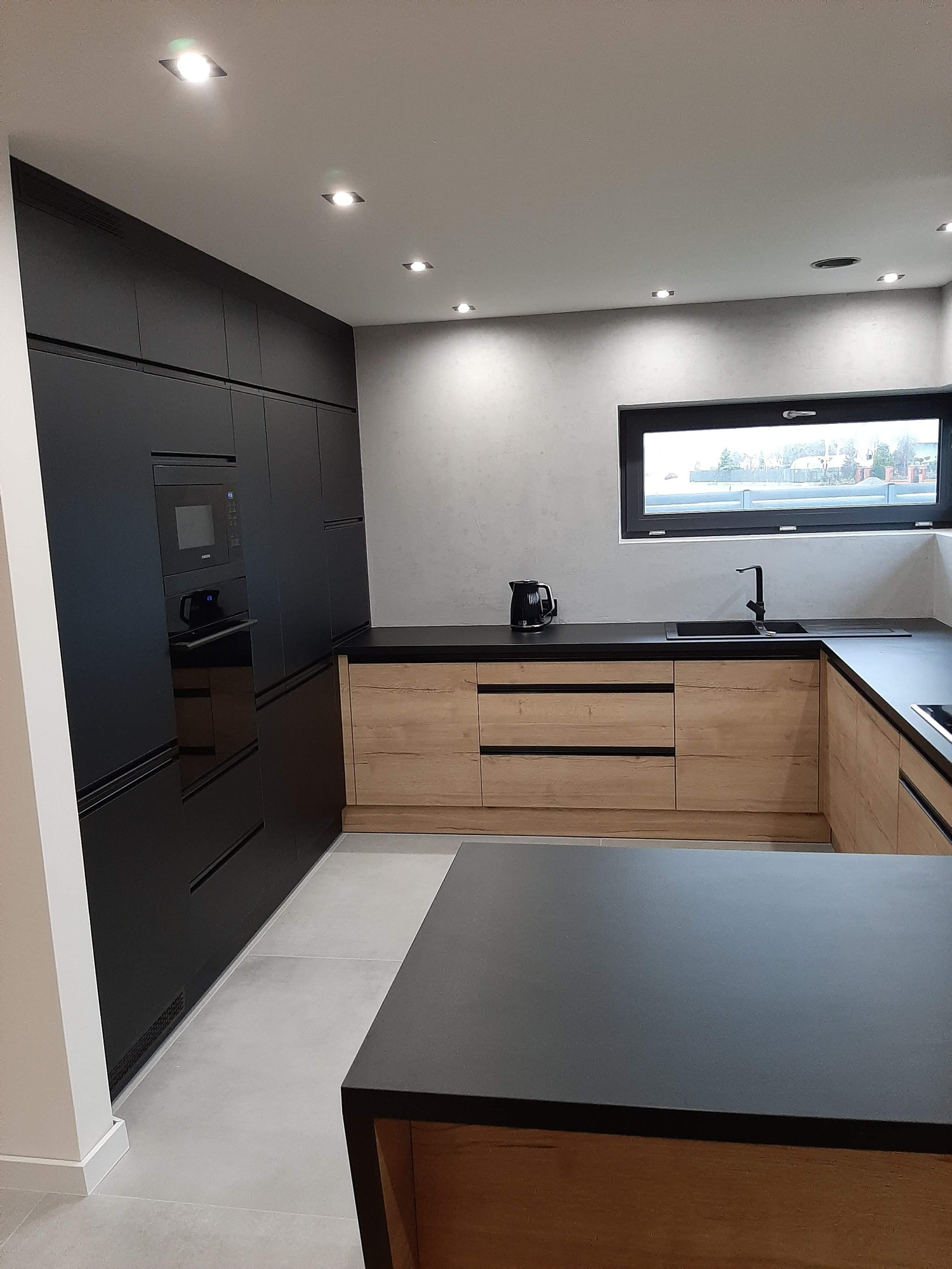 Nowoczesna kuchnia z czarnymi szafkami wysokimi i drewnianymi szafkami dolnymi, czarnym blatem, oknem z widokiem na zewnątrz i minimalistycznym oświetleniem punktowym.