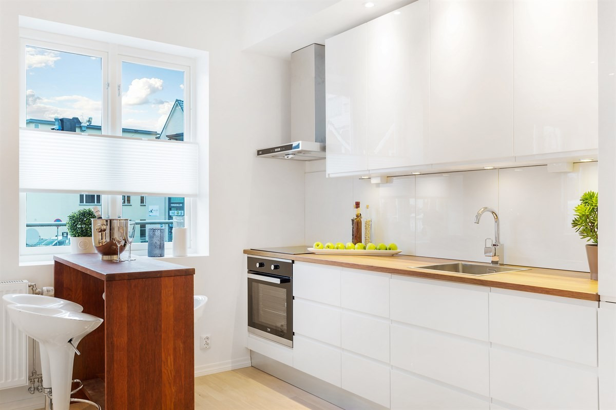 Jasna kuchnia z białymi, lakierowanymi szafkami i drewnianym blatem, uzupełniona o bar z ciemnego drewna i wysokie krzesła barowe.